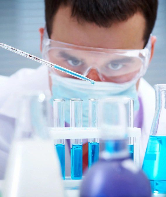 A man in lab coat and goggles looking at blue liquid.